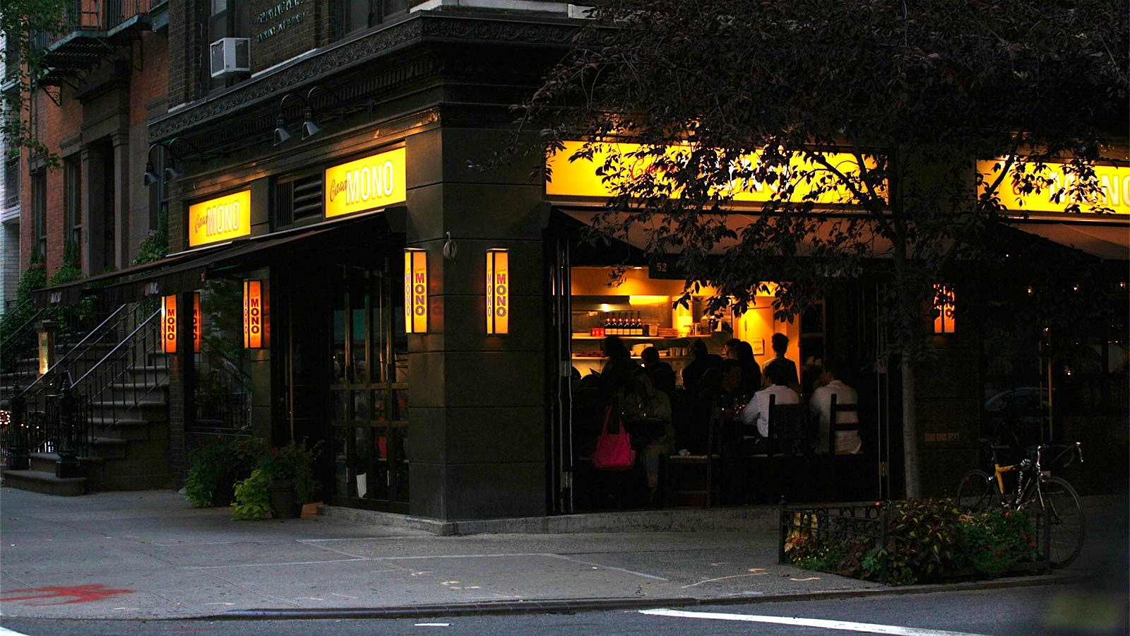  People eating in Casa Mono’s dining room, viewed from outside through large windows beneath yellow Casa Mono signs