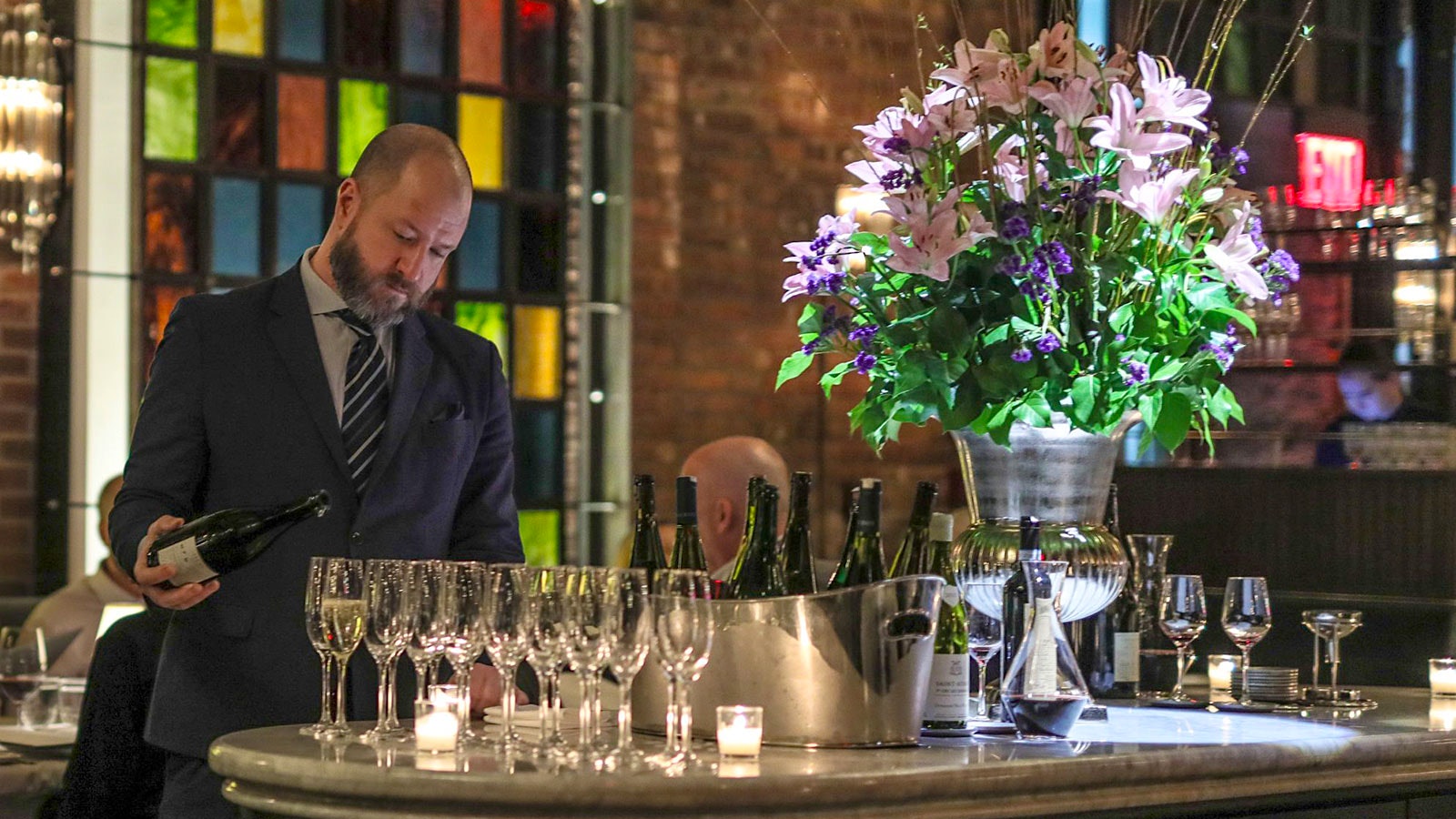  A member of the Temple Court restaurant team pouring sparkling wine into stemmed wineglasses on the same table as a silver vase of flowers, with brick walls behind them and a view of a window made of green, blue and red glass panels