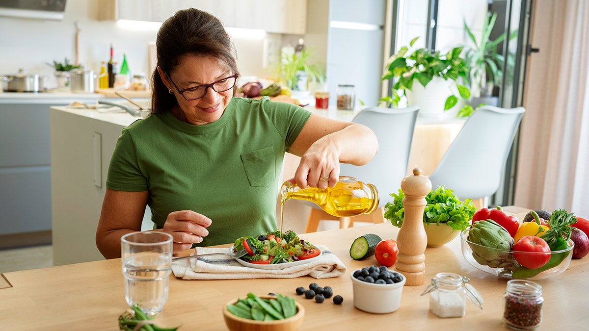 A smiling woman wearing a green shirt pours olive oil on her salad.