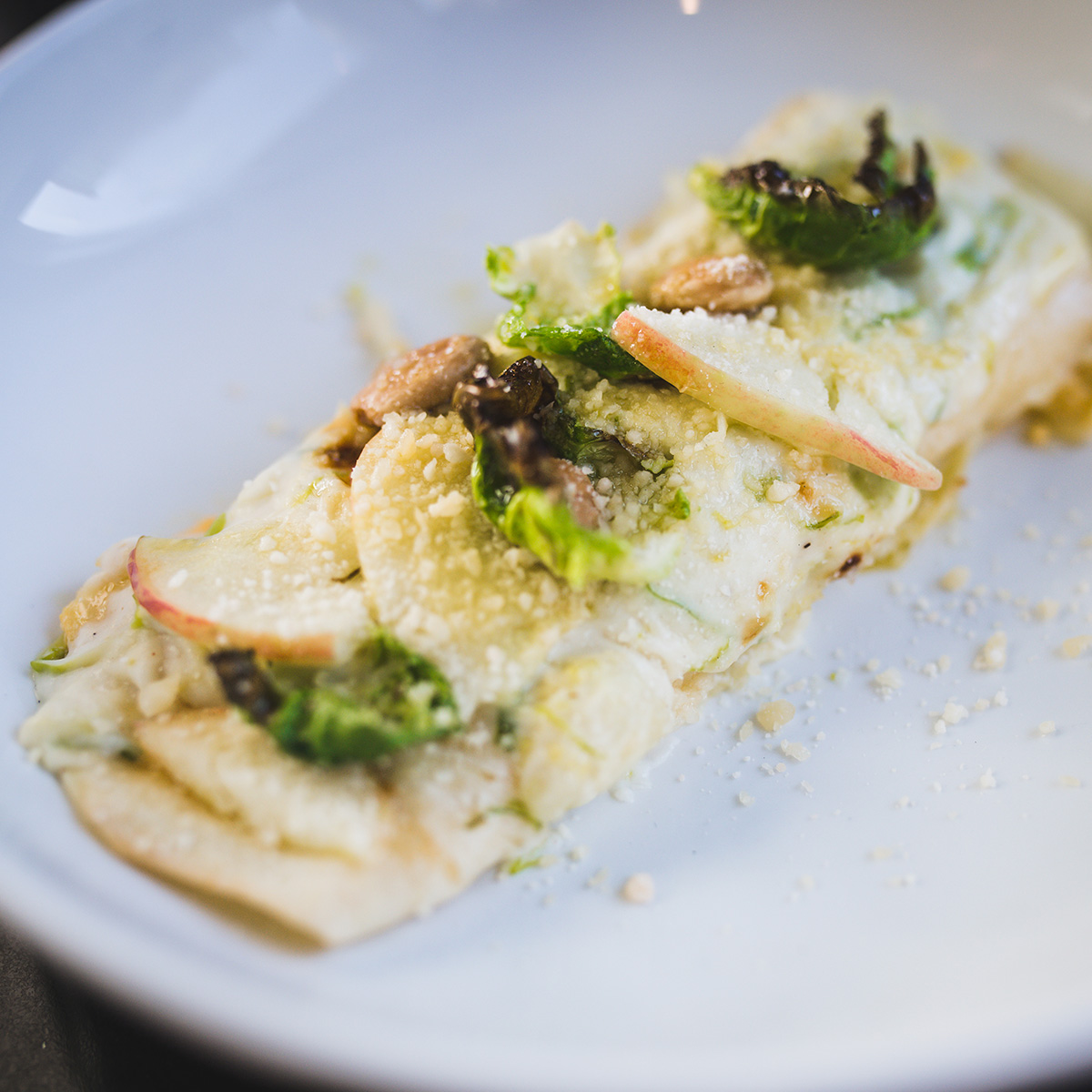 Photo: A large piece of butternut squash manicotti topped with Brussels sprouts, pickled apple, and almonds is shown on a white plate. A slight sprinkling of Parmesan cheese tops the dish.
