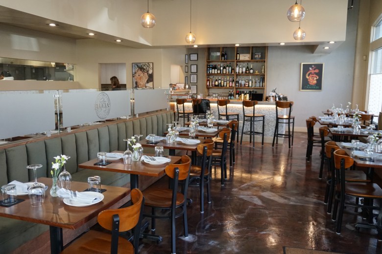 Trattoria Lucia restaurant dining room with green banquette seating, wood chairs, set tables, open kitchen, and full bar.