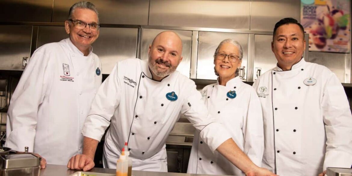 Four chefs in white uniforms smiling in a kitchen. They stand closely together, showing a sense of camaraderie. The background includes stainless steel appliances and countertops.
