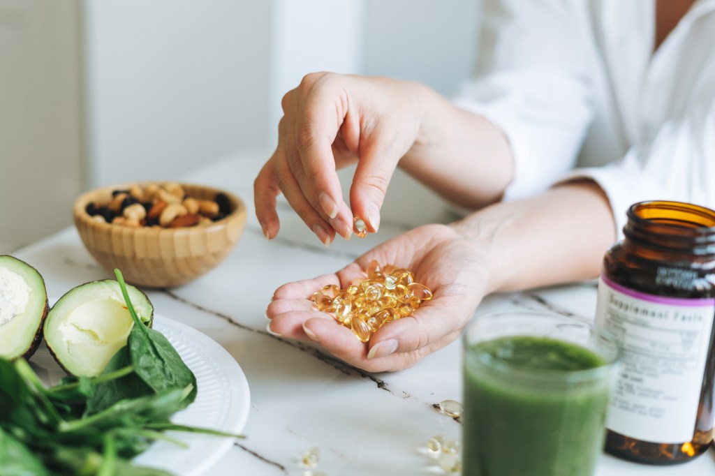 A woman's hands holding Omega-3 and Vitamin D capsules, with green vegetables and nuts on a counter, and a bottle of supplements.