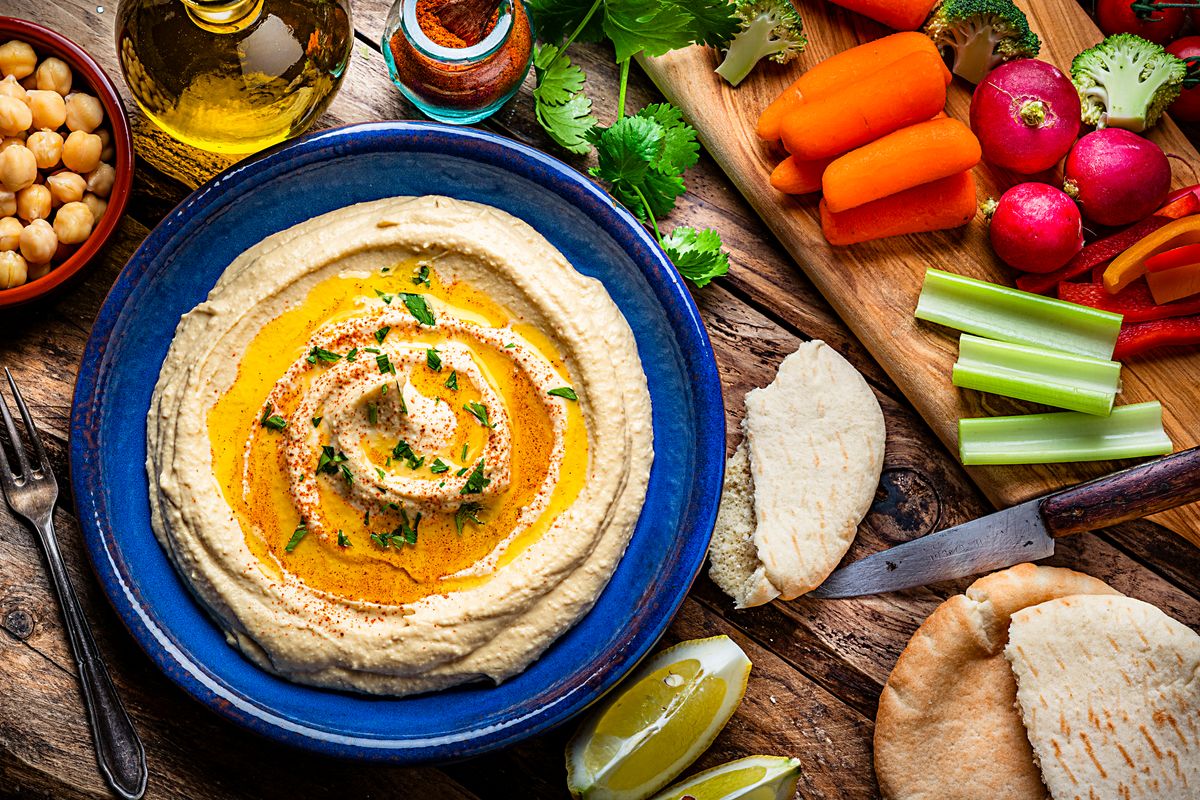 Vegan food: top view of a blue plate with hummus and olive oil shot on rustic wooden table. Pita bread and some fresh vegetables like radish, celery, baby carrots and broccoli are around the plate and complete the composition. High resolution 42Mp studio digital capture taken with Sony A7rII and Sony FE 90mm f2.8 macro G OSS lens