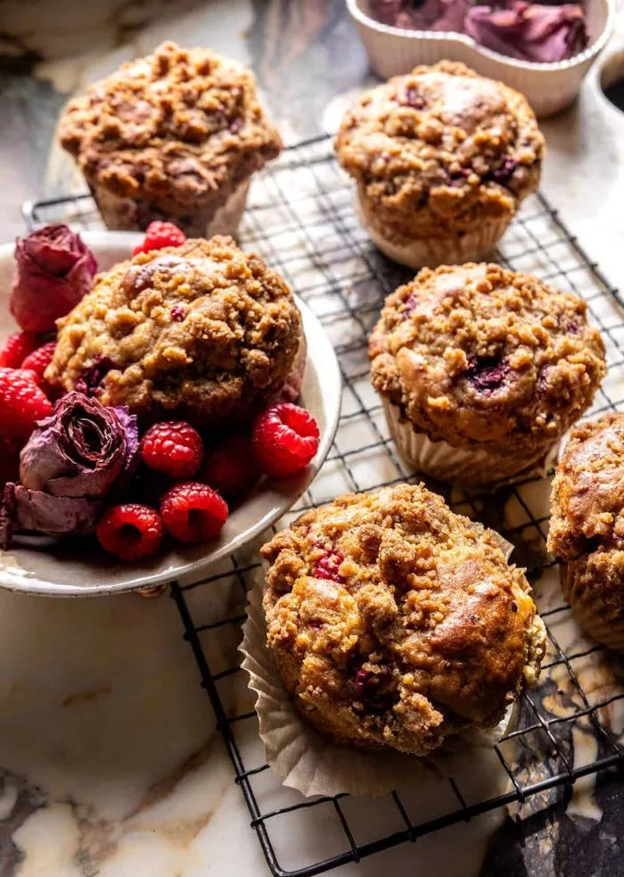 Muffins with crumb topping on a cooling rack next to a bowl of raspberries and dried rose petals on a marble surface