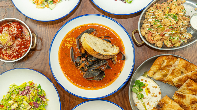 An overhead flat lay of a variety of Italian dishes arranged on a wooden table. The central dish is a white bowl with a blue rim containing mussels in a tomato broth. Other visible dishes include meatballs in sauce, fried calamari, flatbread with a white dip, and several plates of fresh chopped salads.