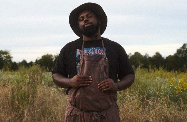 A confident Black man with a full beard and a dark bucket hat stands in a field of tall grass and wildflowers. He is wearing a black t-shirt with a “Boyz n the Hood” graphic and a brown apron. He looks directly at the camera with a neutral expression, holding his hands at his waist. The background consists of trees and an overcast sky.