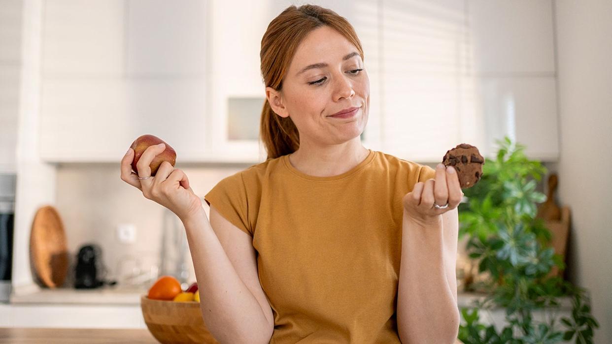 Young woman deciding what to eat between a red apple and a chocolate cookie, holding them in her hands, sitting in her modern kitchen, struggling with a healthy lifestyle.