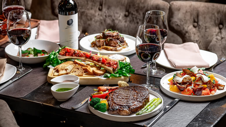 Steak, other dishes, and glasses of red wine on a dark wooden table