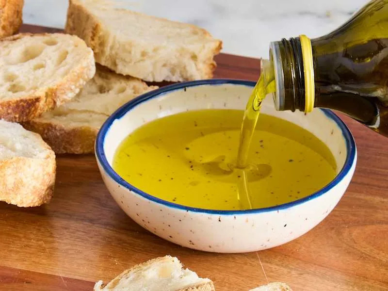 Sliced bread on a wooden board with a bowl of olive oil being poured into it.