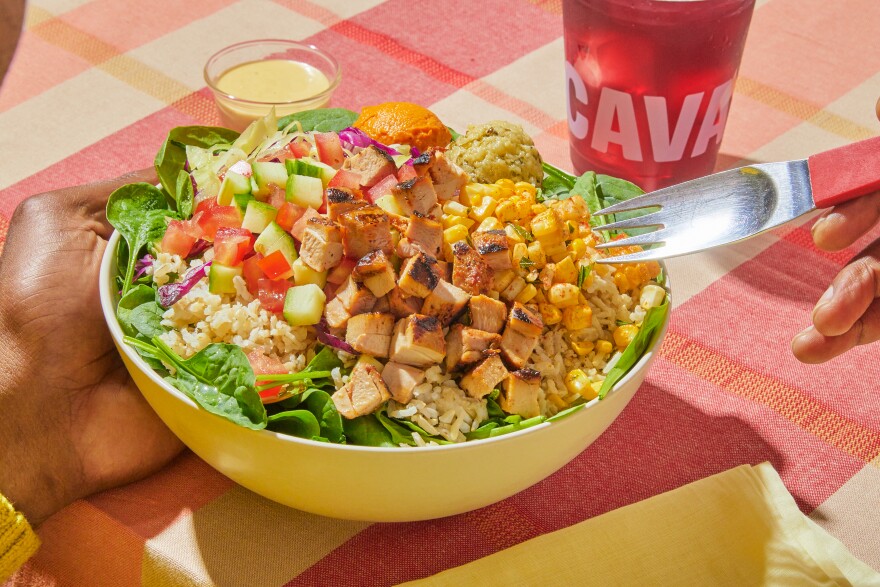 A hand holds a bowl filled with salad greens, rice, chicken and other toppings with a fork hovering overhead.