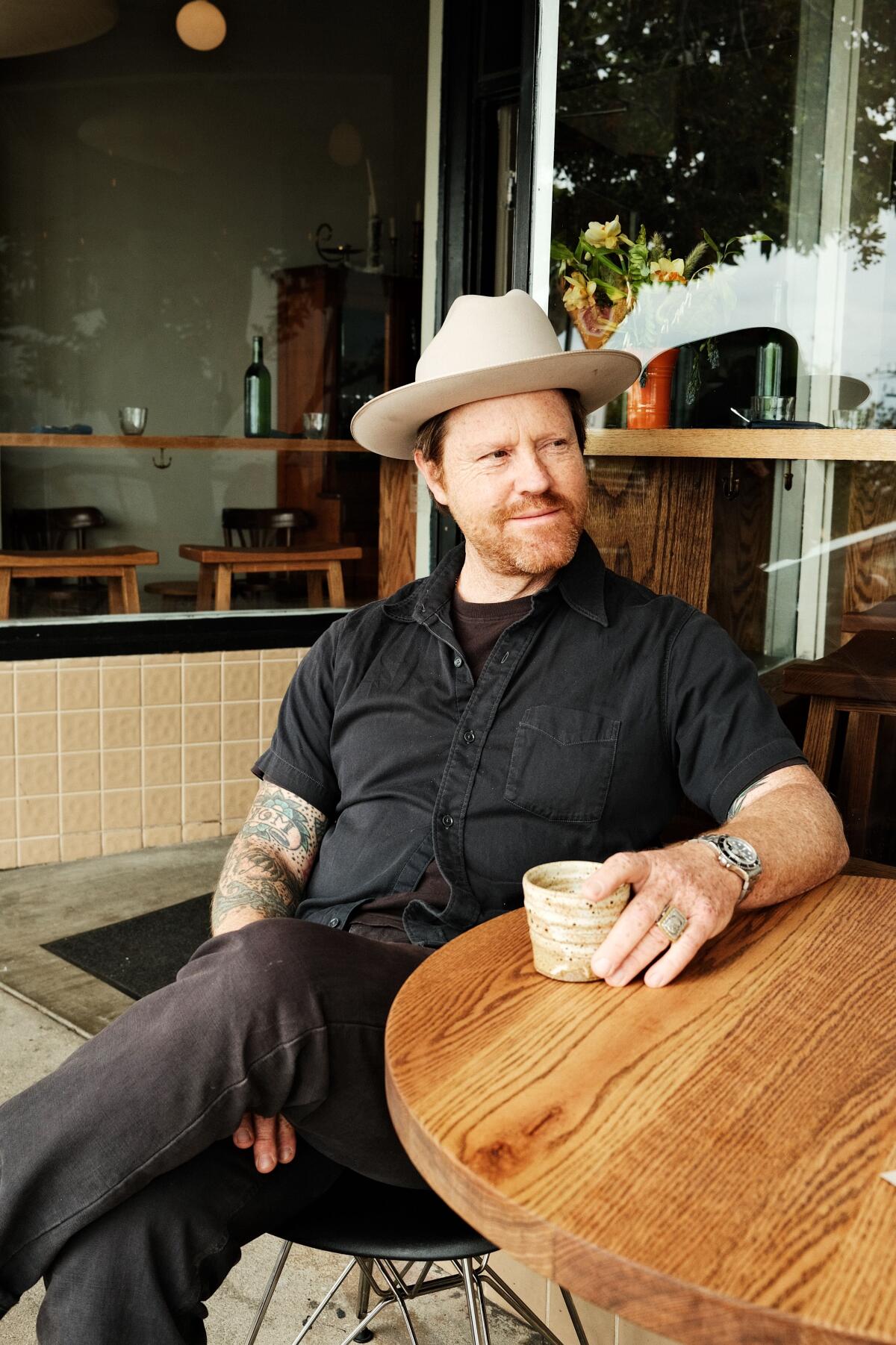 A man sits holding a ceramic mug of coffee at a wood table 