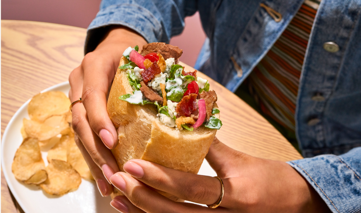 Hand holding a sandwich with various fillings and a plate of chips.