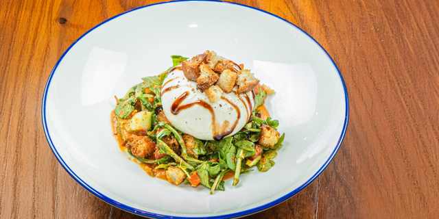 An overhead, close-up shot of a white plate holding a Burrata salad. The large ball of fresh white cheese, drizzled with balsamic glaze, sits atop a mix of greens, tomatoes, and croutons, resting on a wooden table.