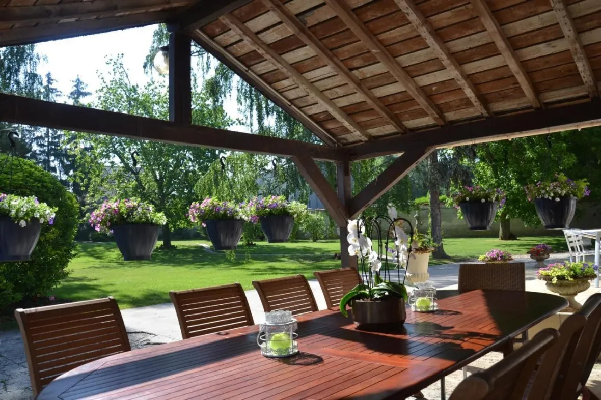 Outdoor dining area with a wooden table, chairs, and potted flowers, overlooking a green lawn.