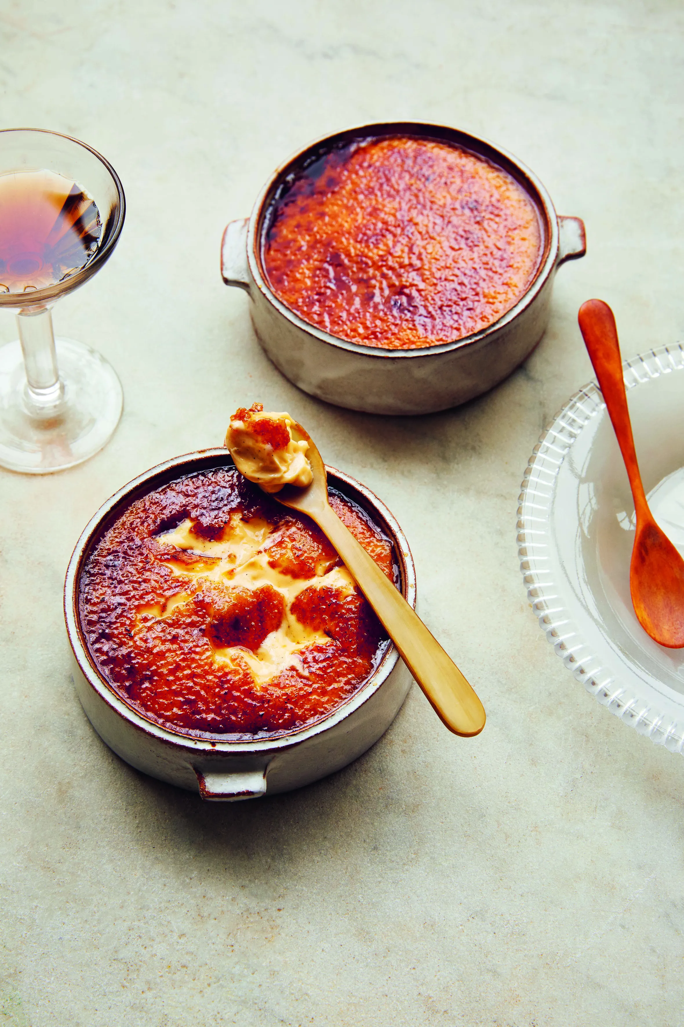 Two creme brûlées with a wooden spoon holding a scoop of creme brûlée, a glass of amber liquid, and an empty plate.