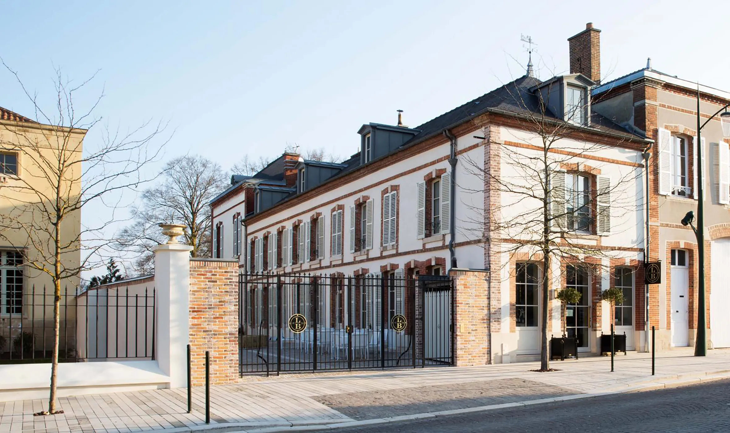 The exterior of Le 25bis by Leclerc Briant, a building with white walls, red brick detailing, and dark gray shutters, seen from across the street.