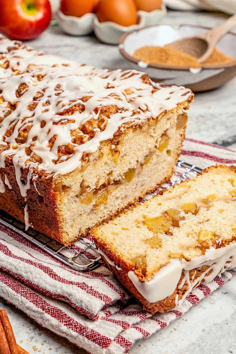 Iced apple cinnamon loaf with a slice cut, showing chunks of apple and cinnamon swirls, on a cooling rack over a striped cloth