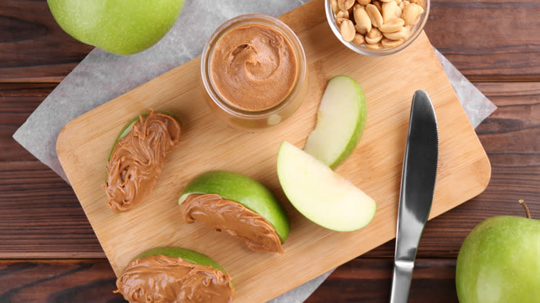 Wooden cutting board with glass jar of peanut butter in the center and Granny Smith apple slices around it with peanut butter on them