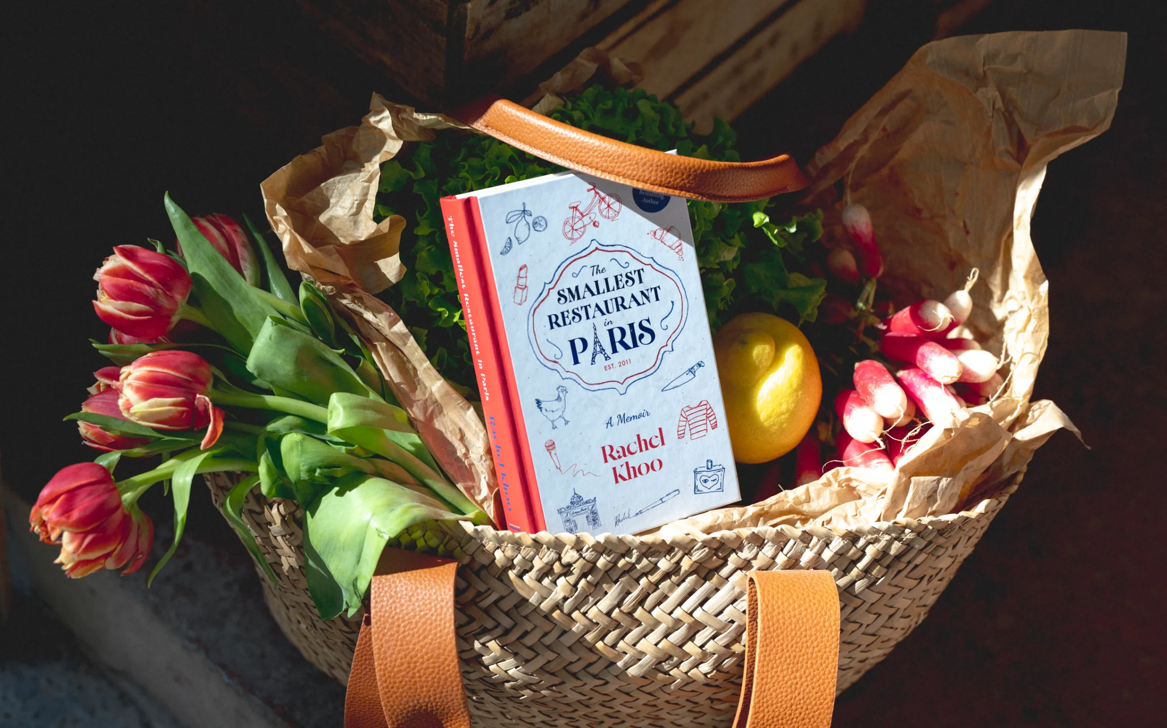 A wicker bag filled with groceries, including red tulips, lettuce, radishes, a lemon, and a book titled "The Smallest Restaurant in Paris" by Rachel Khoo.