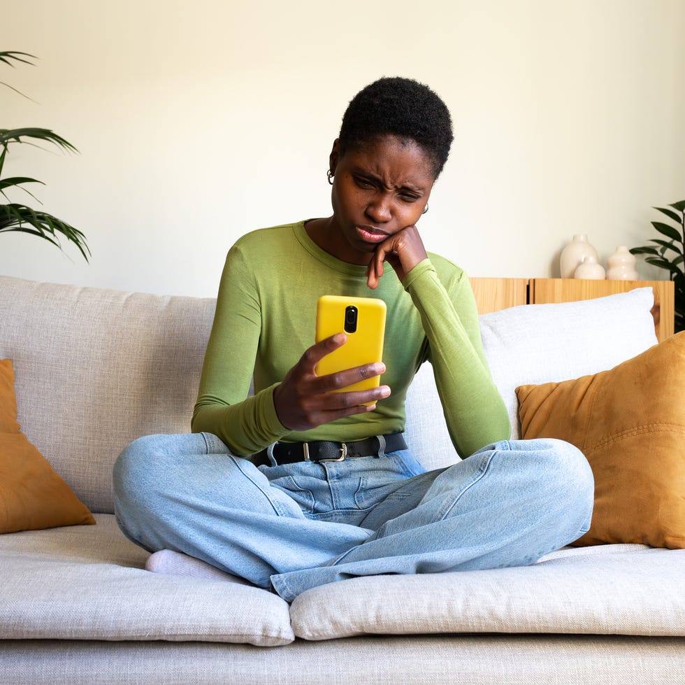 young woman sitting on a sofa and looking at a smartphone with a confused expression
