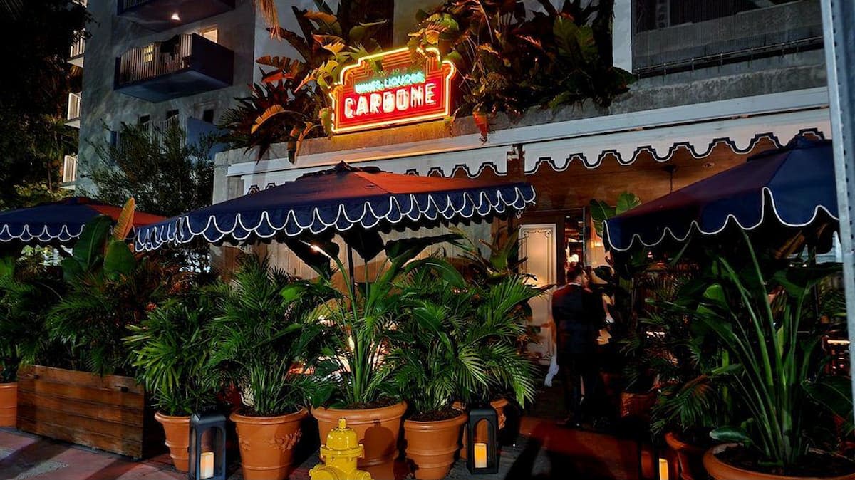 A warmly lit restaurant with a neon “CARBONE” sign above the entrance, reminiscent of iconic Italian chain restaurants in America, features outdoor seating under blue awnings and lush green plants in pots lining the patio at night.