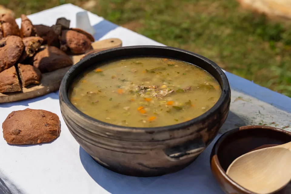 A rustic bowl of hearty soup with vegetables on a table, accompanied by a wooden spoon and fresh bread