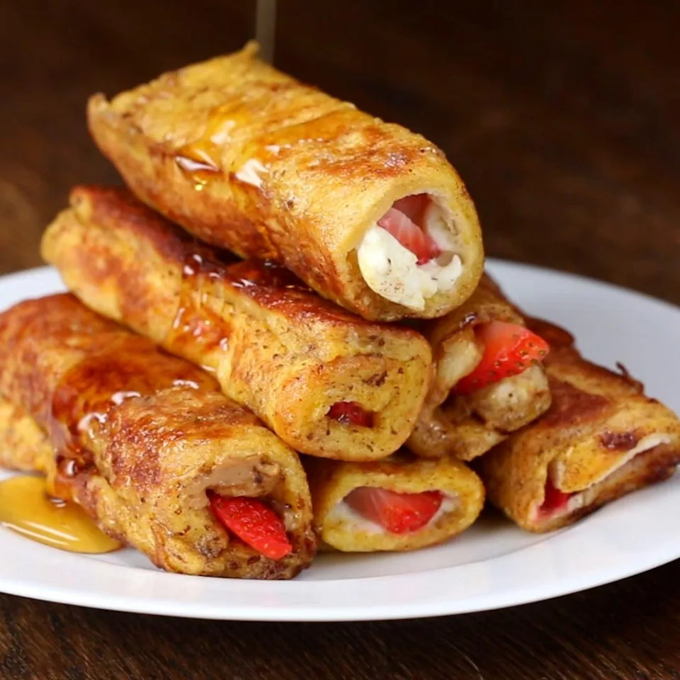 Stack of French toast roll-ups with strawberries and cream cheese filling, drizzled with syrup, on a white plate