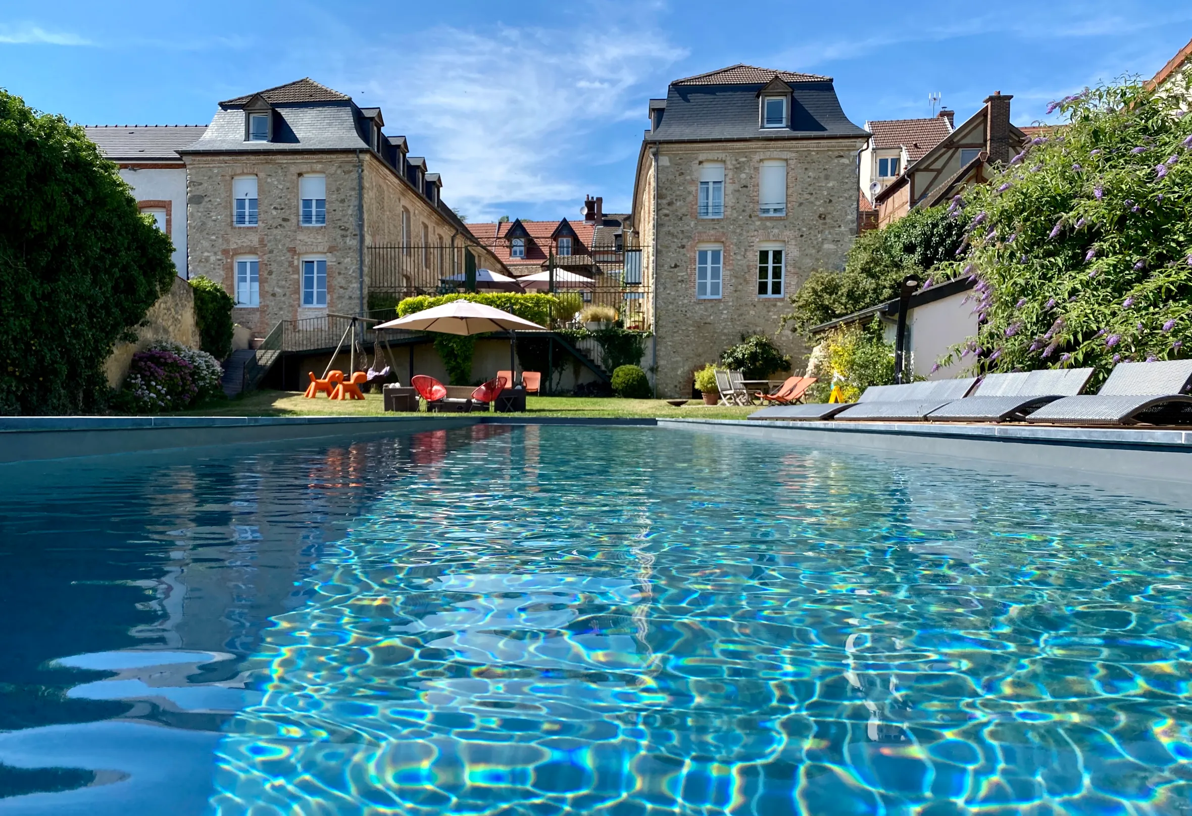 A sparkling blue swimming pool with two stone buildings and green gardens in the background.