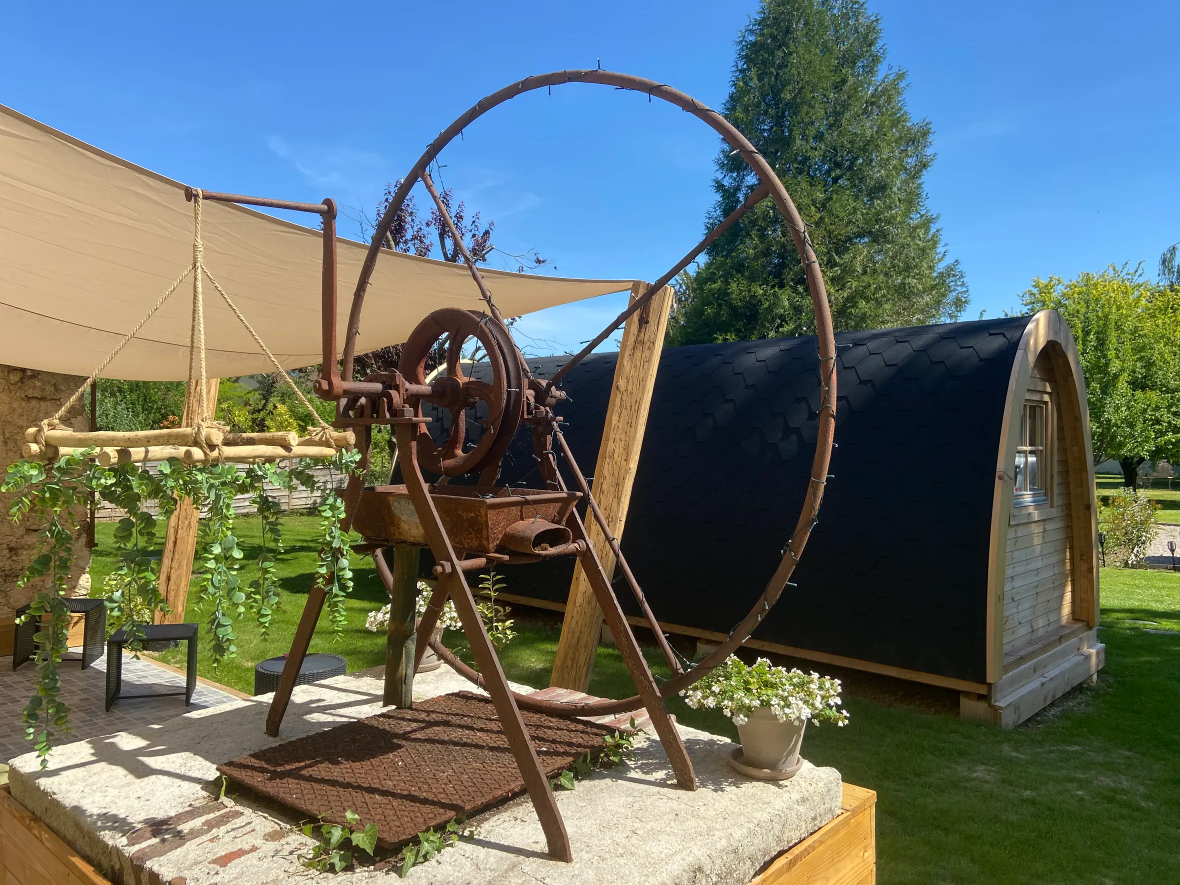 A large, rusted metal wheel structure with a small potted plant in front and a wooden cabin with a black roof behind, under a blue sky.