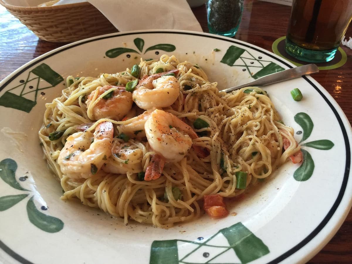 A plate of shrimp scampi pasta with angel hair noodles, shrimp, diced tomatoes, and green onions, garnished with black pepper—just like you'd find at Italian chain restaurants in America—served on a decorative plate with a fork.