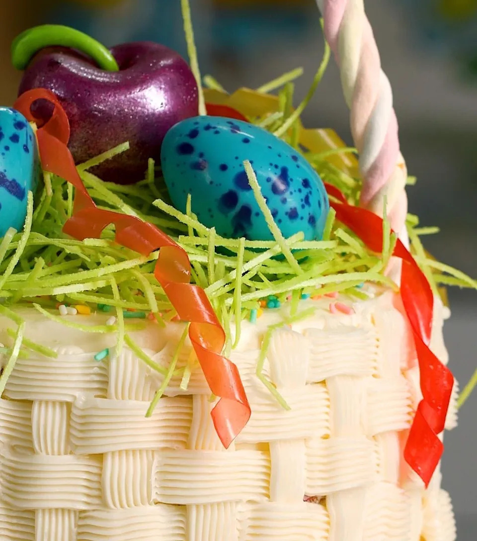 Decorative cake styled like a woven basket, topped with candy eggs and an apple, and garnished with colorful sugar decorations