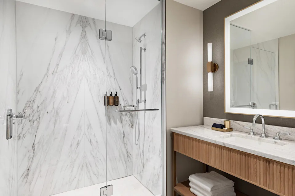 A walk-in shower with white marble walls. A sink with a large mirror is in the foreground.