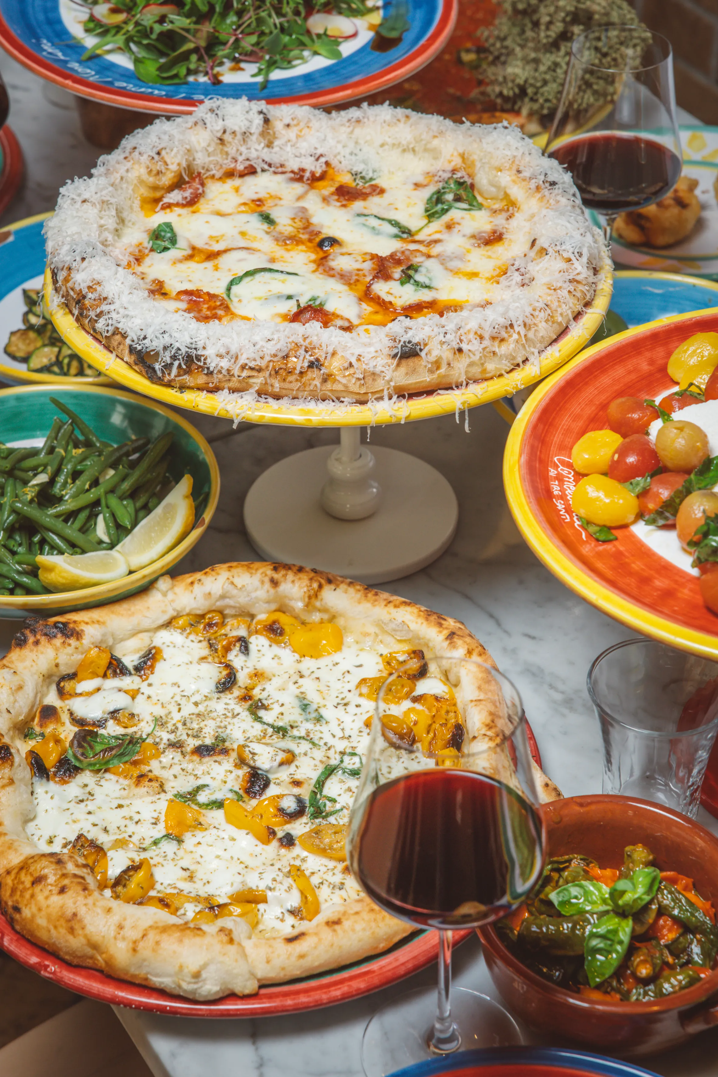 Two pizzas, a glass of red wine, and a variety of colorful side dishes, including a bowl of green beans with lemon slices.