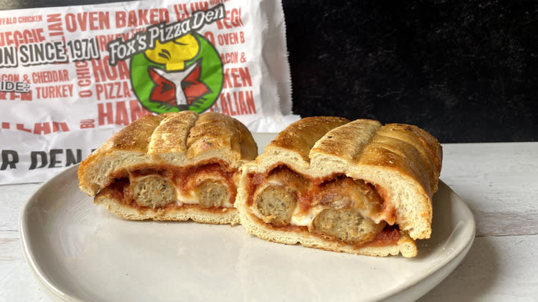 A Fox's Pizza Den meatball sub sliced in half on a plate in front of the to-go packaging