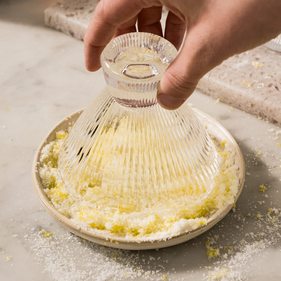A hand holding a glass dish over a plate with salt and lemon zest.