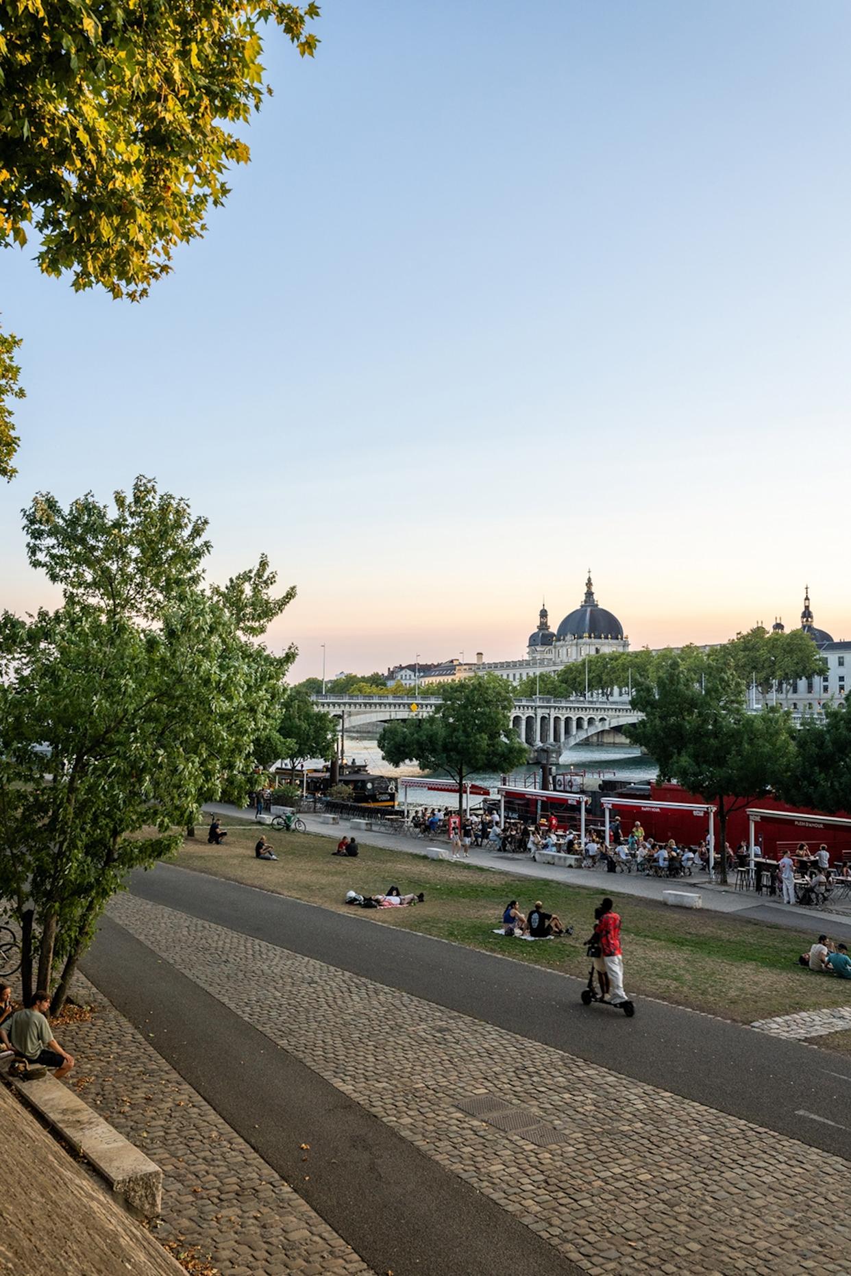 A riverside promenade in a city with outdoor seating and canal boats along the water banks.