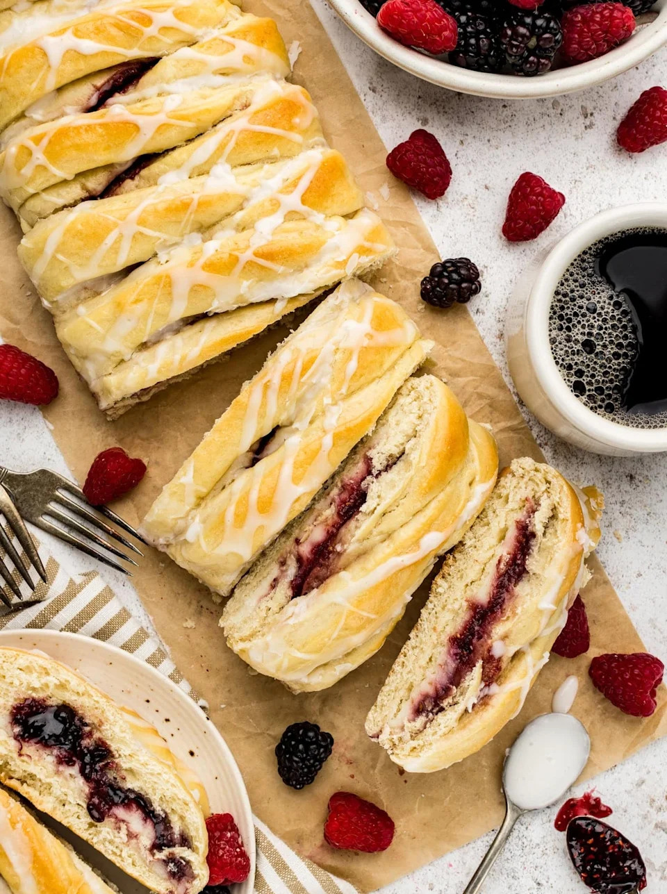 Sliced braided pastry with berry filling and icing, surrounded by fresh berries and a cup of coffee