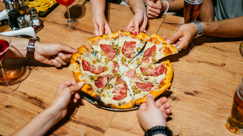 Hands reaching toward a pizza and pulling out slices