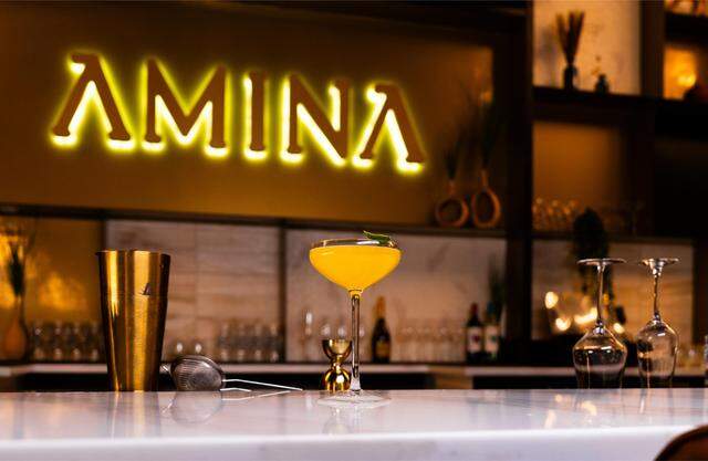 A bright yellow cocktail in a coupe glass is garnished with a lime twist and sits on a white marble bar top. In the background, a warm, glowing “AMINA” sign illuminates the bar area.