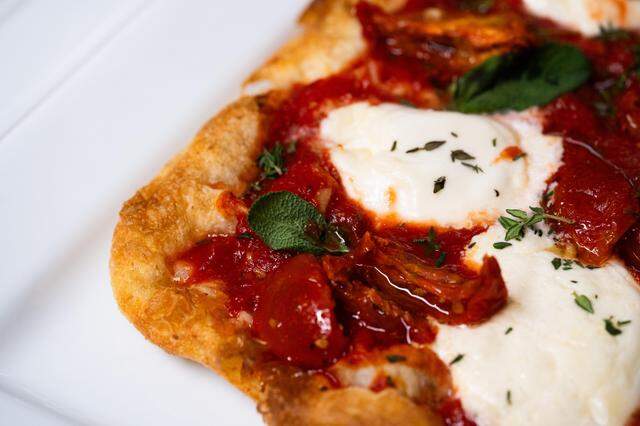 A close-up shot of a rustic flatbread features vibrant red tomato sauce topped with melted white cheese and fresh green herbs. The crust is golden brown and crispy, served on a white rectangular plate.