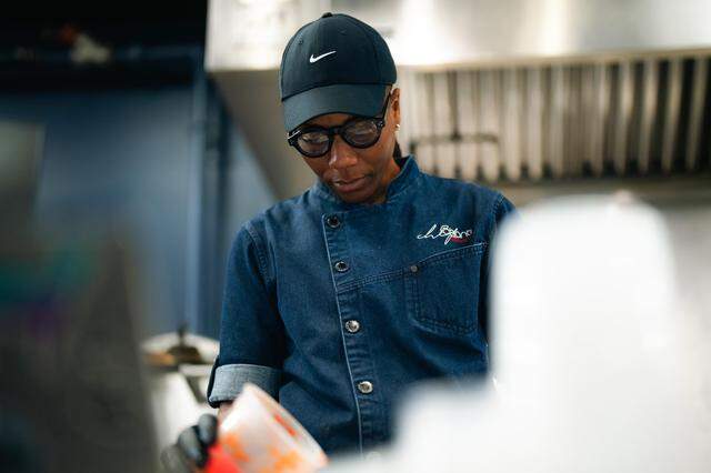 A chef wearing a denim jacket, black Nike cap, and glasses focused intently on preparing food in a professional kitchen. The background shows stainless steel kitchen equipment under bright industrial lighting.