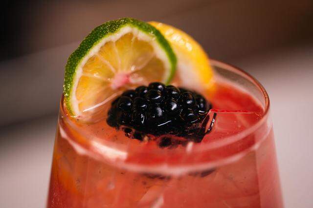 This close-up captures a refreshing pink cocktail over ice, garnished with a large blackberry and slices of lime and lemon. The drink is served in a clear glass, highlighting the condensation and vibrant fruit colors.