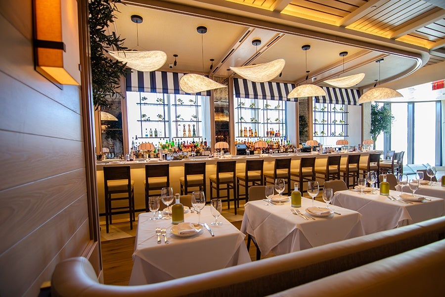 A modern restaurant interior featuring a long bar with high wooden stools and a variety of bottles displayed on shelves behind it. In front of the bar, there are several tables covered with white tablecloths, each set with plates, glasses, cutlery, and small olive oil bottles. The ceiling has unique, curved pendant lights, and large windows with striped awnings allow natural light to fill the space. The overall atmosphere is bright and inviting.