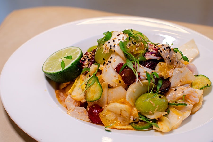 A colorful salad featuring sliced cucumbers, green and red grapes, and leafy greens, garnished with sesame seeds and microgreens, served with a halved lime on a white plate.