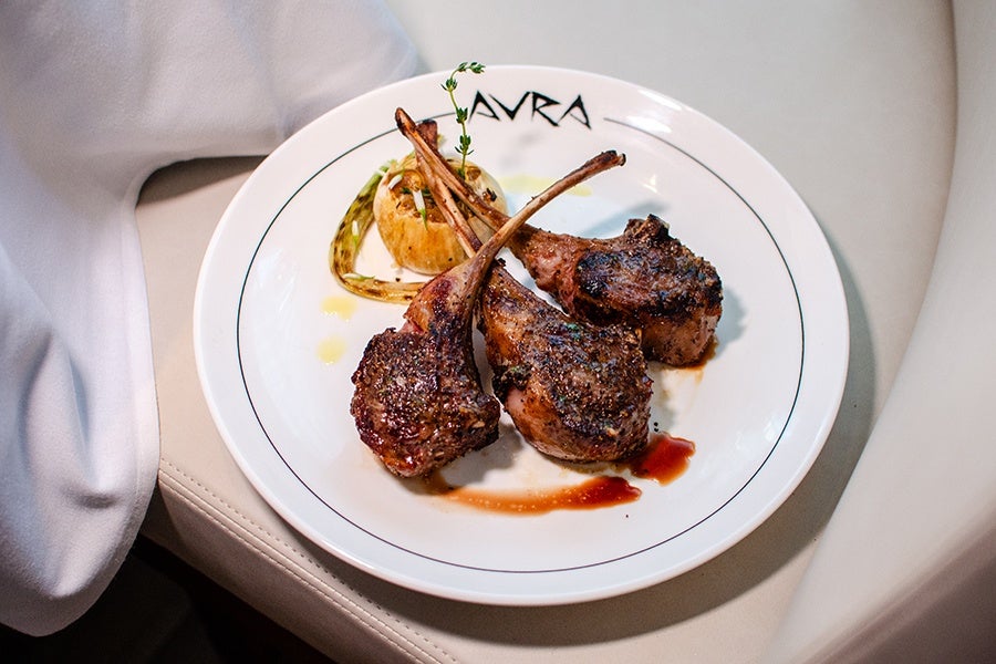 Three grilled lamb chops seasoned with herbs are served on a white plate with a roasted garlic bulb and a drizzle of sauce, garnished with a sprig of thyme. The plate has the word 
