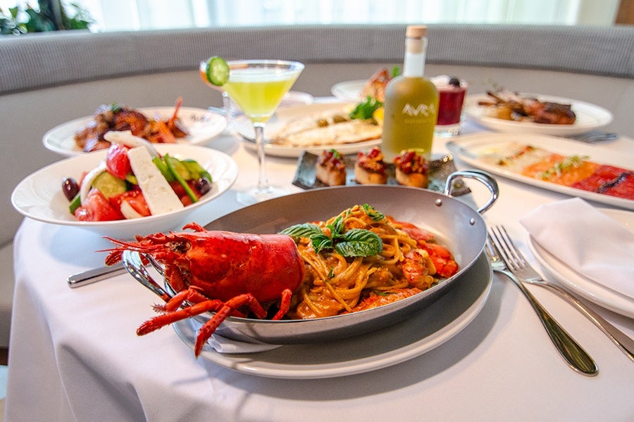 A plated lobster pasta dish garnished with fresh basil in a metal pan on a white tablecloth. Surrounding the main dish are various other plates including a Greek salad with feta cheese, a cocktail with a cucumber slice, bruschetta, grilled meat, and a bottle of olive oil. Silverware and a folded white napkin are placed to the right of the main dish.