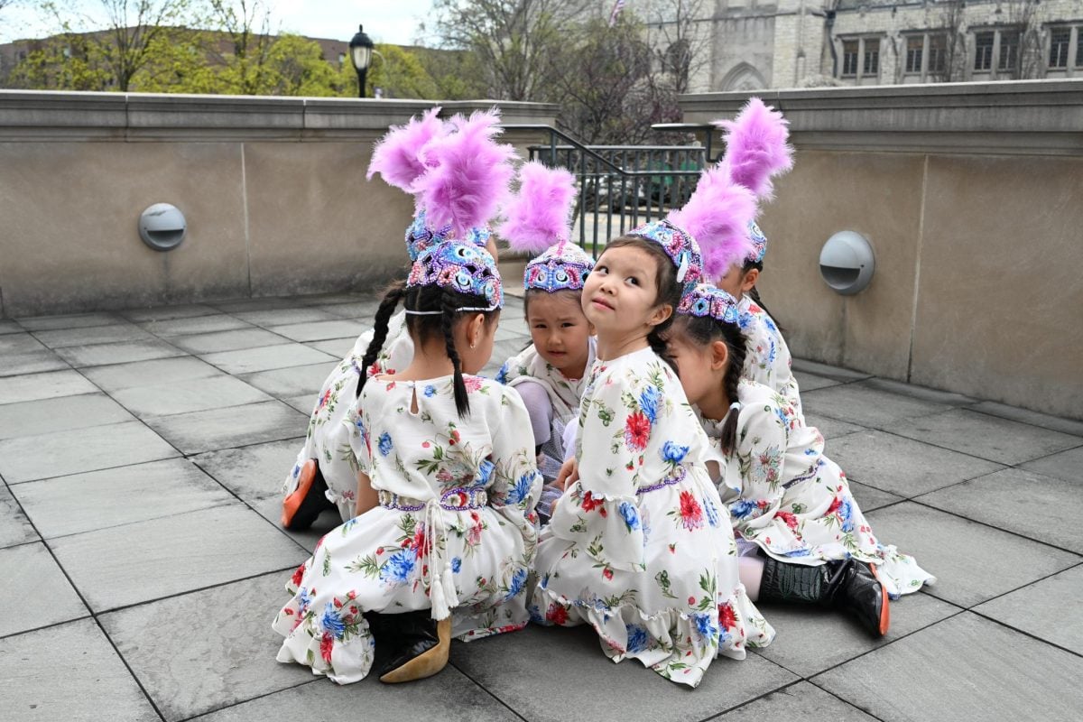 Children wearing white dresses and hats with purple feathers sitting on the ground in a circle. 