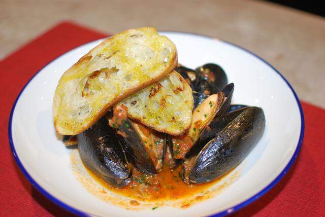 A high-angle, close-up shot of steamed mussels served in an orange-red tomato broth in a white bowl with a blue rim. The dish is generously topped with two slices of toasted, oiled bread (crostini) and rests on a bright red placemat.