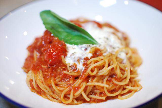 A tight, close-up shot of a plate of spaghetti covered in a chunky red tomato sauce. The dish is topped with creamy melted cheese and garnished with a large, fresh green basil leaf.